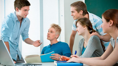 Fünf Schülerinnen und Schüler befinden sich an einem Tisch im Klassenzimmer. Alle Blicke sind auf eine Person gerichtet.