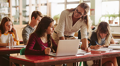 Eine Schülerin sitzt an einem Laptop. Die Lehrerin steht daneben und unterhält sich mit ihr.