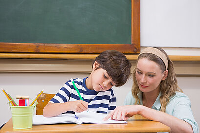 Ein Schüler schreibt in ein Heft. Die Lehrerin zeigt in das Heft.