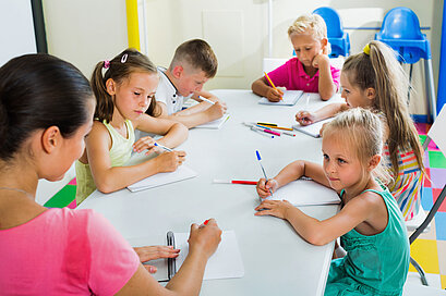 Eine Lehrerin sitzt mit mehreren jungen Schülerinnen und Schülern an einem Gruppentisch und schreiben.