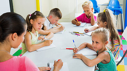 Eine Lehrerin sitzt mit mehreren jungen Schülerinnen und Schülern an einem Gruppentisch und schreiben.