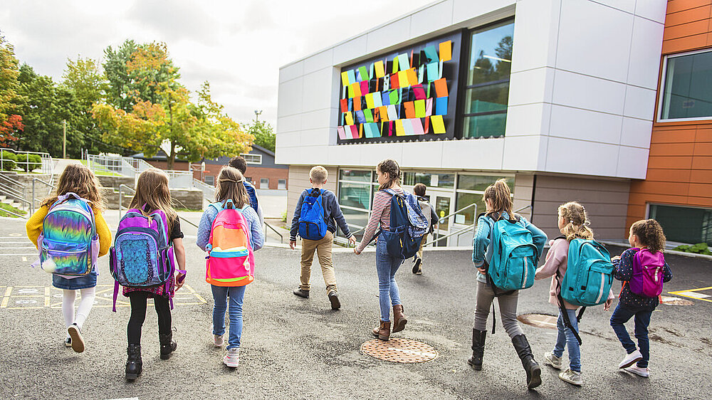 Mehrere Kinder gehen mit ihren Schulränzen auf dem Rücken vor einem Schulgebäude.