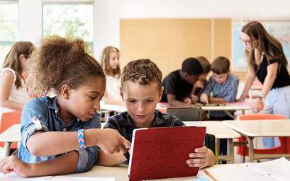 In einem Klassenzimmer arbeiten zwei Kinder gemeinsam an einem Tablet. Im Hintergrund sind weitere Kinder und die Lehrerin zu sehen.