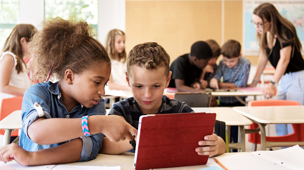 In einem Klassenzimmer arbeiten zwei Kinder gemeinsam an einem Tablet. Im Hintergrund sind weitere Kinder und die Lehrerin zu sehen.