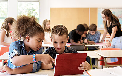 In einem Klassenzimmer arbeiten zwei Kinder gemeinsam an einem Tablet. Im Hintergrund sind weitere Kinder und die Lehrerin zu sehen.